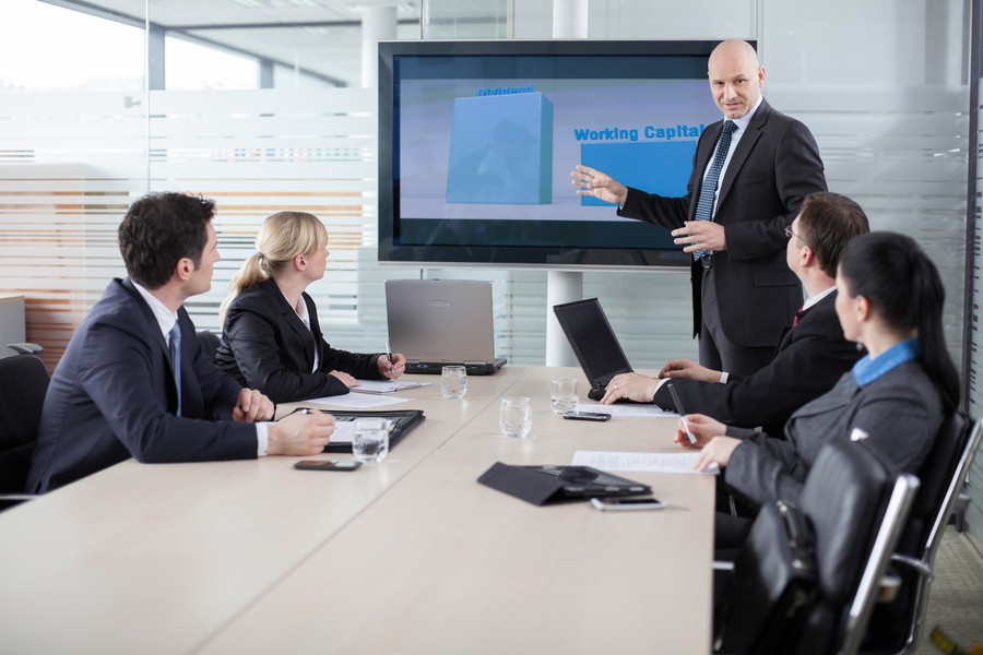 A man presents a business presentation to colleagues in a board room. There is a high-end display mounted on the wall.