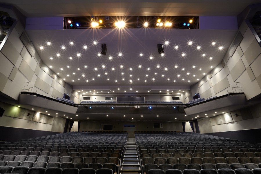 A modern auditorium with tiered seating, acoustic wall panels, and a ceiling adorned with recessed lighting.